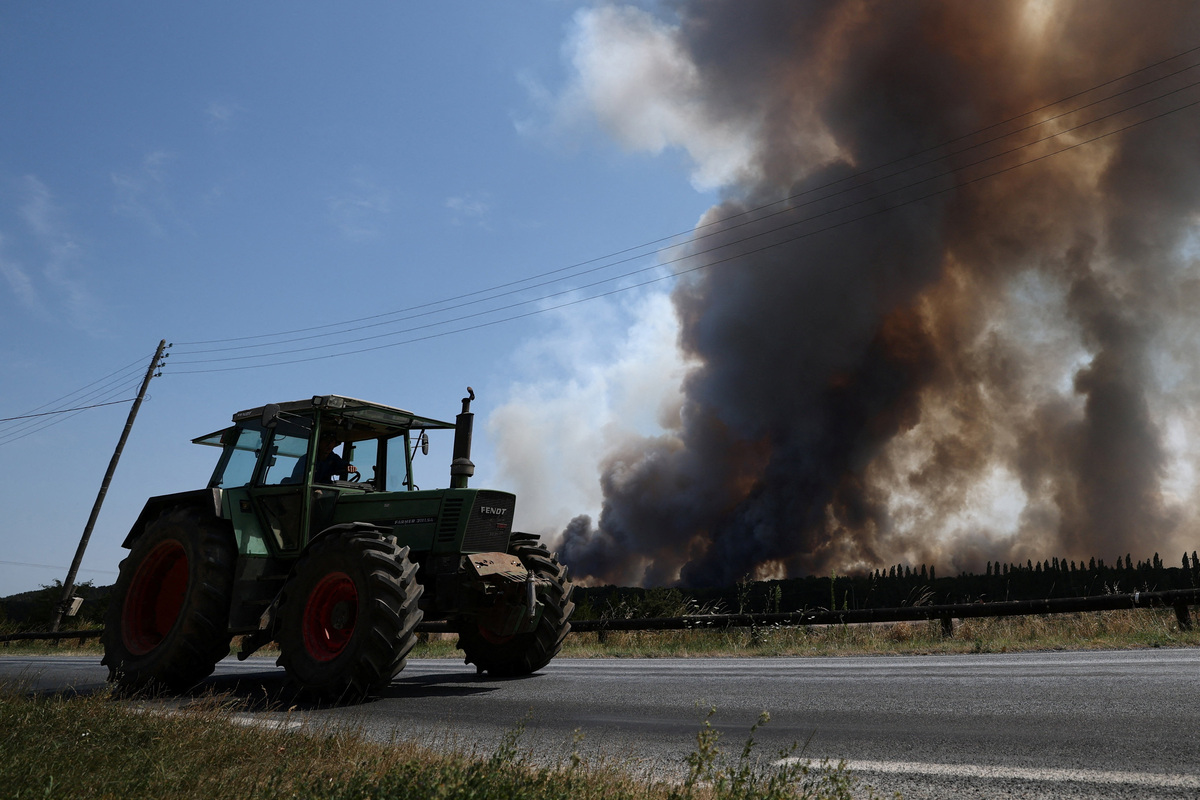 A farmer drives his tractor along a road in Milly-la-Foret as smoke rise from a wildfire in the Fontainebleau forest near Paris during a heatwave affecting large parts of France, July 13, 2026. REUTERS/Stephane Mahe