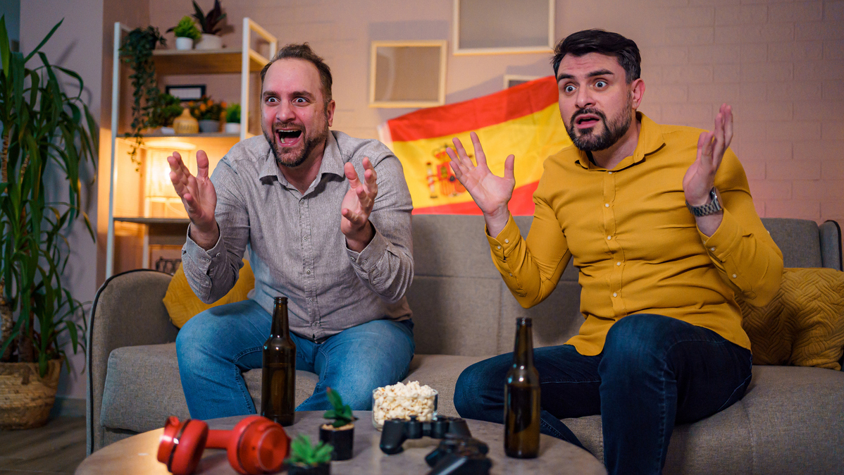 Two friends express excitement while watching a sports event at home, surrounded by snacks.