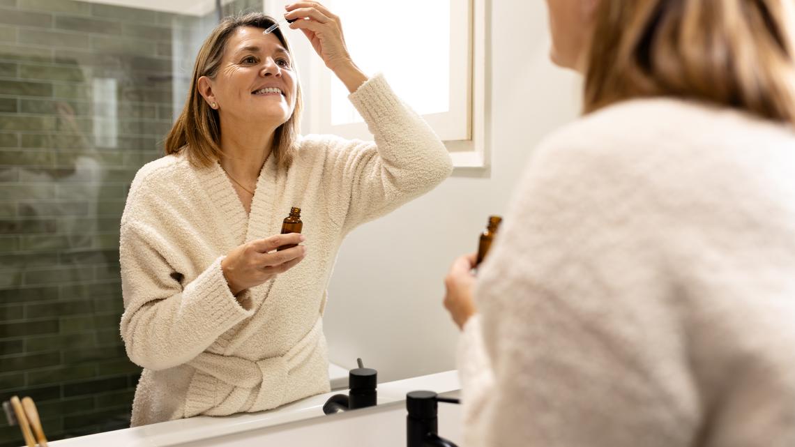 Smiling mature woman applying cosmetic serum on her forehead in the bathroom mirror