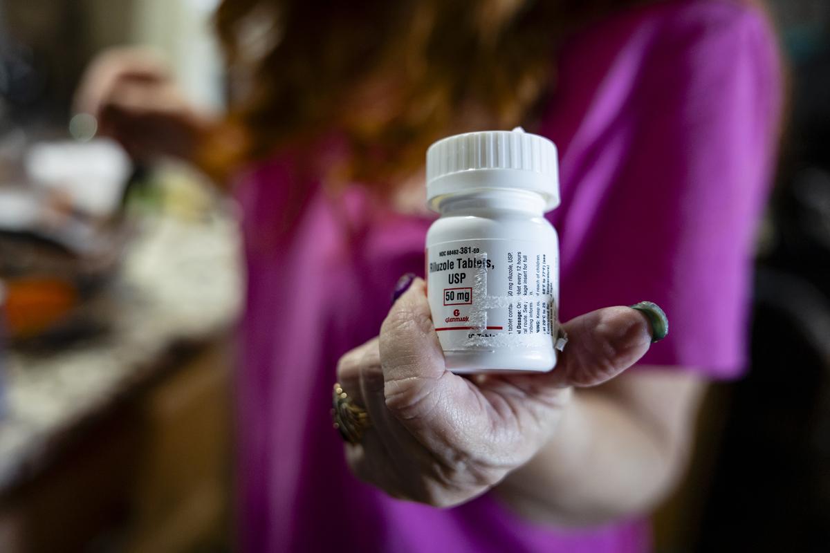 Amanda Sifford holds a bottle of riluzole, one of only two other FDA-approved ALS medications, which can extend survival by several months at her home in Cape Coral, Fla., on Jan. 25, 2026. The drug, tofersen, made by Biogen and marketed as Qalsody, is for a small subset of patients. But evidence that breathing and strength can get better for some of them is remarkable for a paralyzing, usually fatal disease. (Eva Marie Uzcategui/The New York Times)