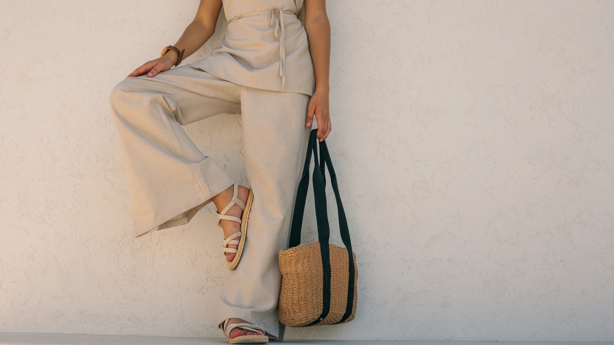 A woman poses in neutral-toned linen clothing, leaning against a textured wall, holding a woven tote bag.