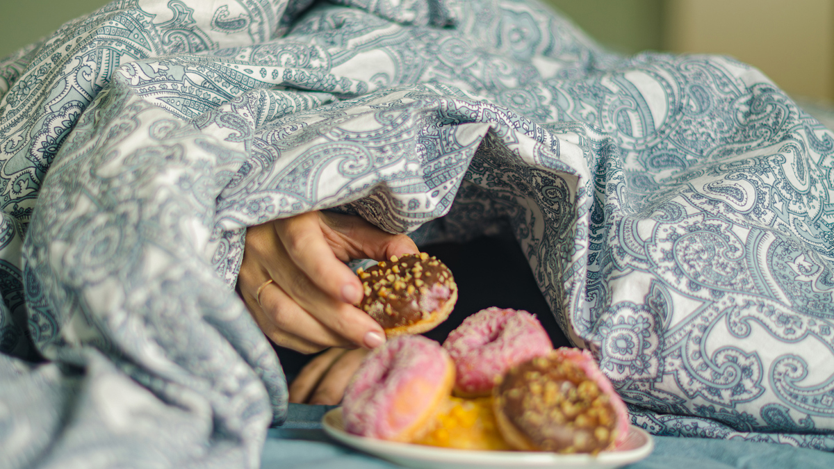 A stock image of a woman reaching hand for a doughnut while lying in bed. 