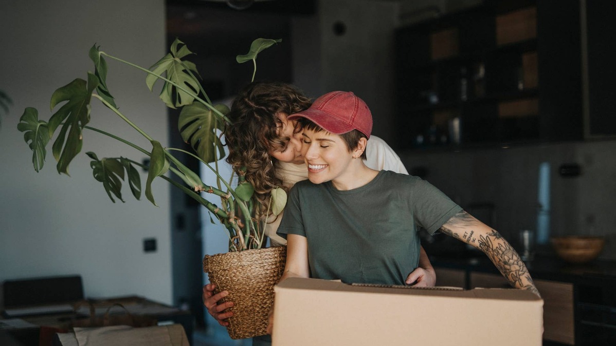 Smiling lesbian woman holding houseplant kissing girlfriend carrying cardboard box during relocation.