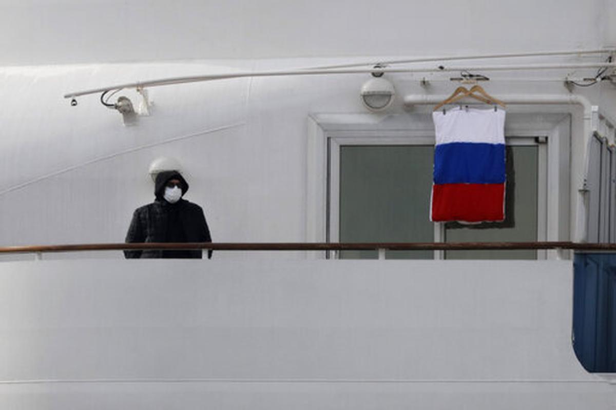 A quarantined passenger on the Diamond Princess cruise ship stands on the balcony of his cabin next to a Russian flag Saturday, Feb. 15, 2020, in Yokohama, near Tokyo. A viral outbreak that began in China has infected more than 67,000 people globally. The World Health Organization has named the illness COVID-19, referring to its origin late last year and the coronavirus that causes it.