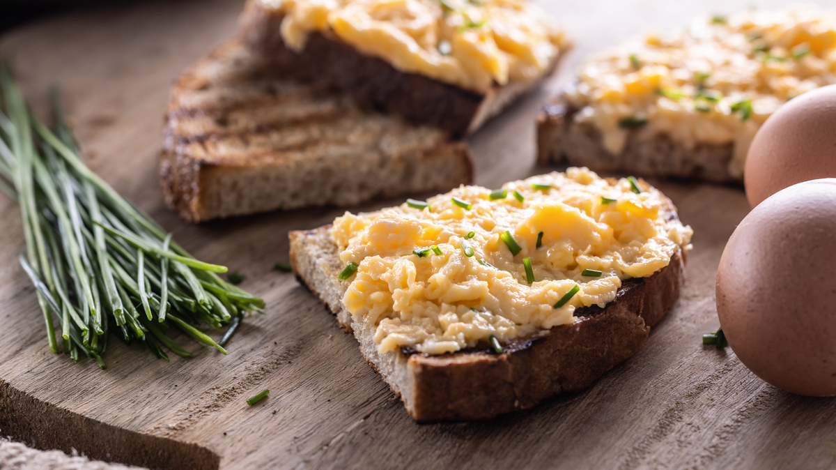 Fresh scrambled eggs spread on a slice of bread with whole eggs and fresh chive on the side.