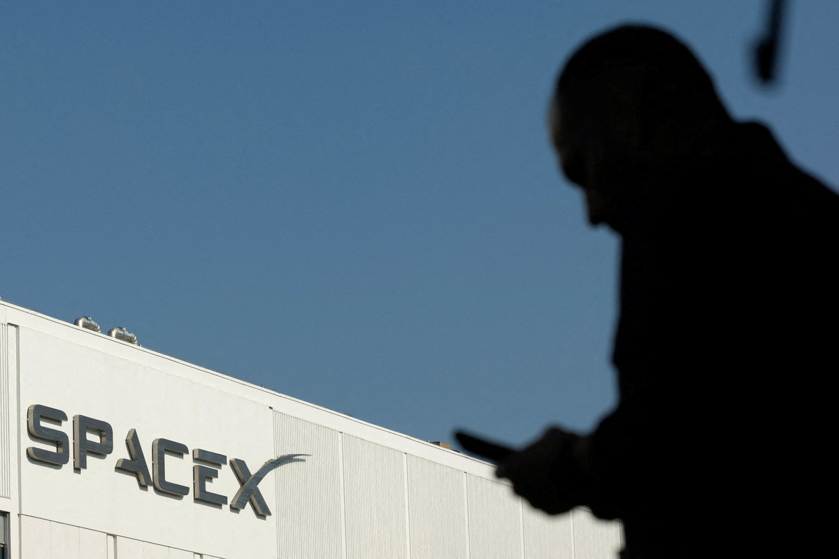FILE PHOTO: SpaceX logo as an employe looks at his phone while making his way to work at the company’s facility on the day of the SpaceX IPO, in Hawthorne, California, U.S. June 12, 2026.  REUTERS/Mike Blake/File Photo