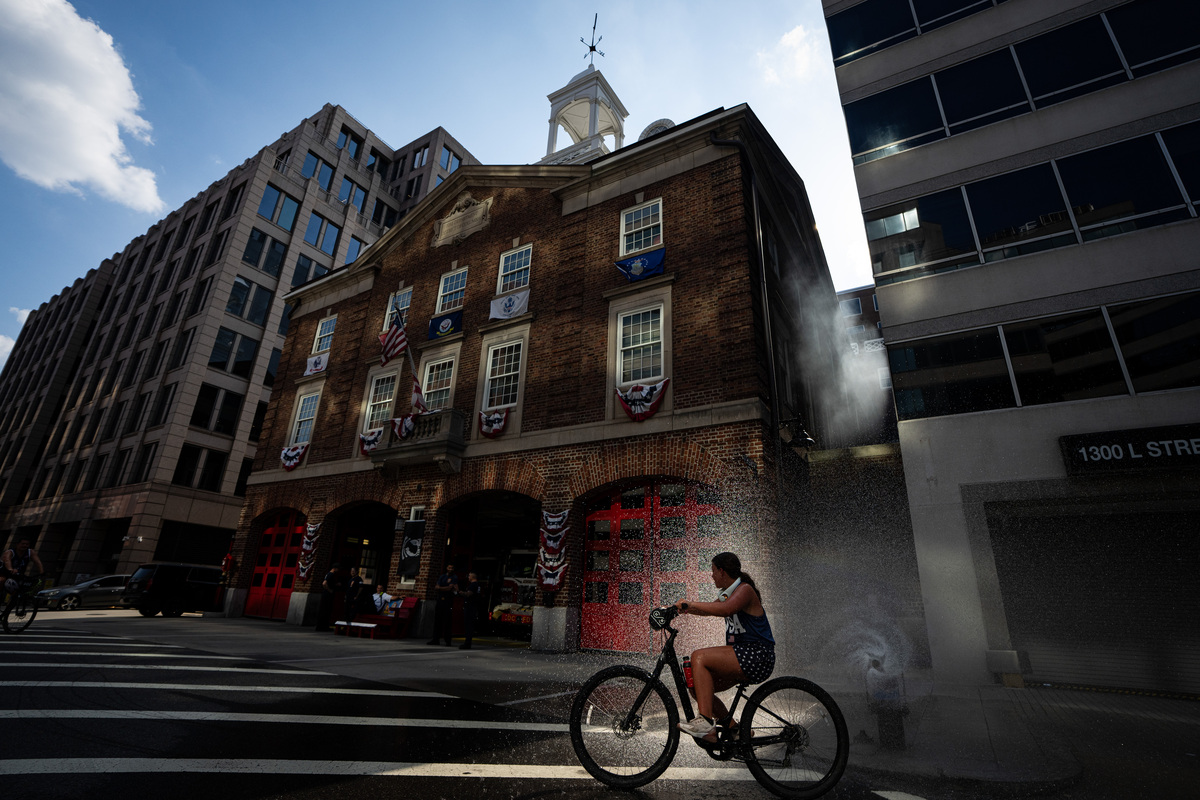 A girl bikes past a fire hydrant spraying water at the Franklin Square DC Fire and EMS station as the temperature hits 99 degrees on July 4, 2026. America celebrates the 250th anniversary of its independence today, with people from one end of the country to the other marking the milestone with fireworks, parades and concerts. (Al Drago/The New York Times)