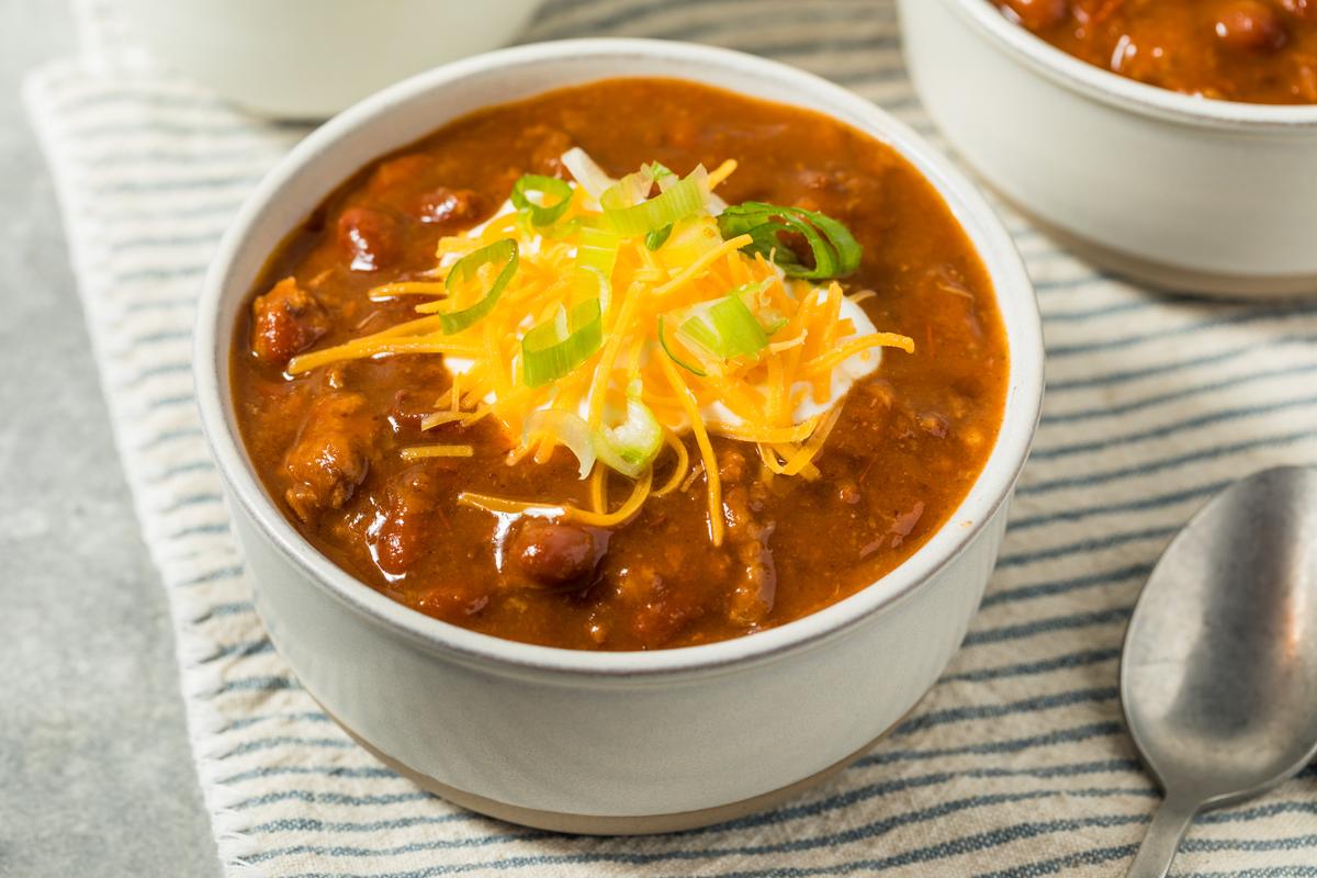 A close-up, high-angle shot of a bowl of chili. The chili has a rich, reddish-brown color with visible beans and ground meat. It is topped with a dollop of white sour cream or yogurt, a mound of shredded yellow and orange cheddar cheese, and a sprinkle of chopped green onions. The bowl sits on a striped blue and white cloth, with a spoon and another bowl partially visible in the background.