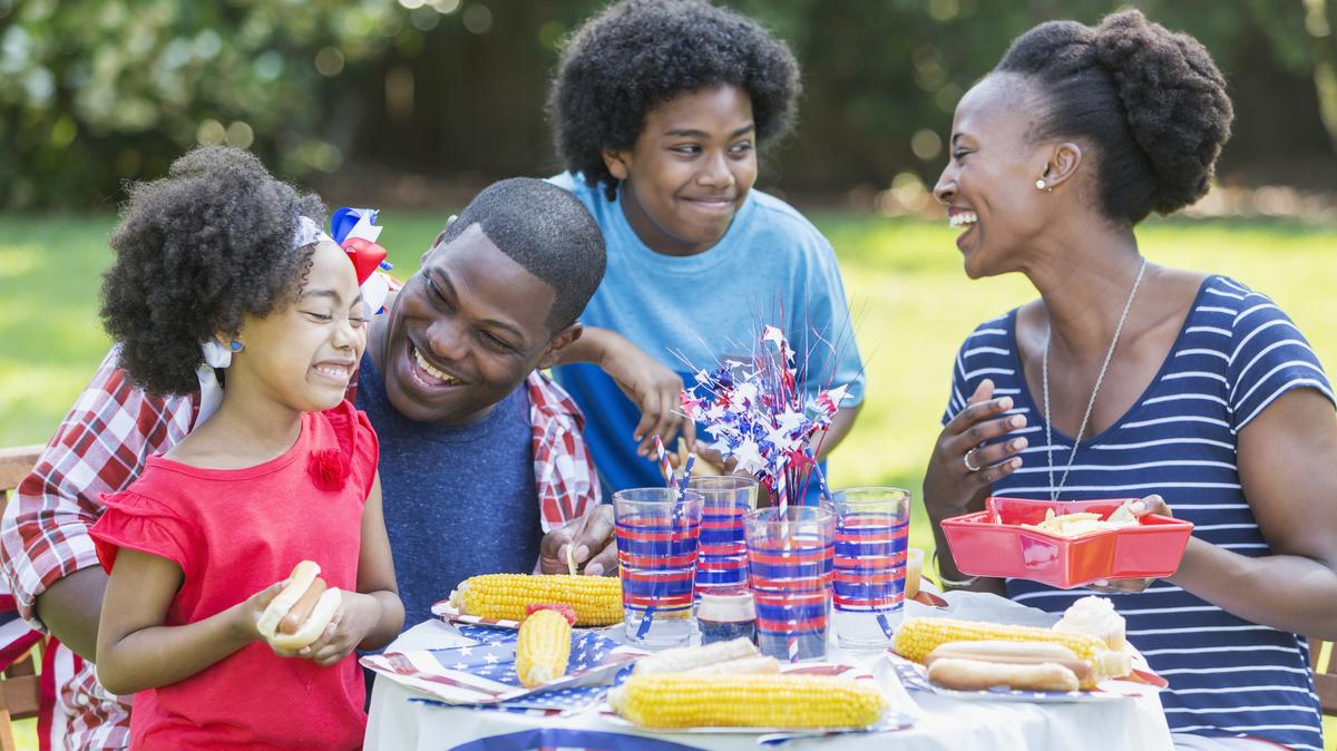 A family enjoying a backyard cookout on the 4th or July or Memorial Day. The children are mixed race African American, Asian and Hispanic. They are eating hotdogs and corn on the cob. The 12 year old boy is standing between his parents, looking at his mother.