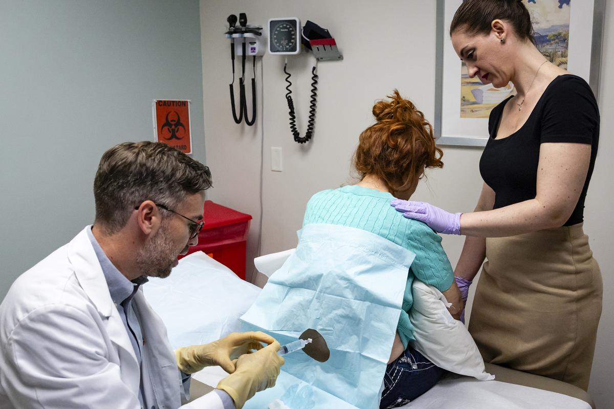 Dr. Nathan Carberry, a neurologist at the University of Miami Health System, administers an infusion of tofersen to Amanda Sifford's spinal canal in Miami on Jan. 9, 2026. The drug, tofersen, made by Biogen and marketed as Qalsody, is for a small subset of patients. But evidence that breathing and strength can get better for some of them is remarkable for a paralyzing, usually fatal disease. (Eva Marie Uzcategui/The New York Times)