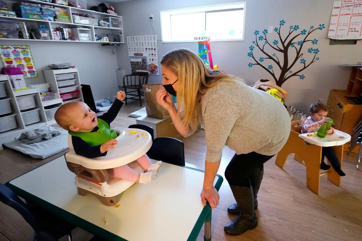 Amy McCoy signs to a baby about food as a toddler finishes lunch behind at her Forever Young Daycare facility, Monday, Oct. 25, 2021, in Mountlake Terrace, Wash. Child care centers once operated under the promise that it would always be there when parents have to work. Now, each teacher resignation, coronavirus exposure, and day care center closure reveals an industry on the brink, with wide-reaching implications for an entire economy’s workforce.