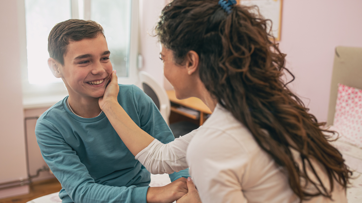 A happy mother touching and cuddling her teenage son on the face in his bedroom. 