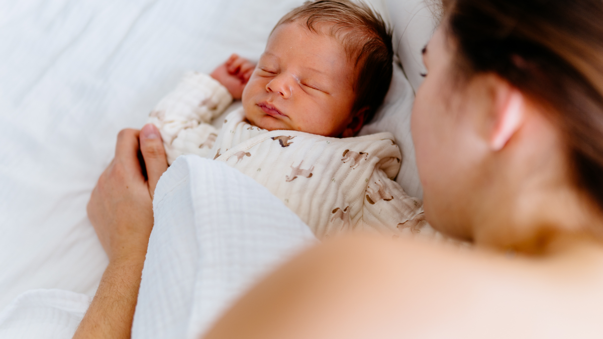 Close-up of newborn baby peacefully sleeping next to mother, wrapped in a soft white blanket. 