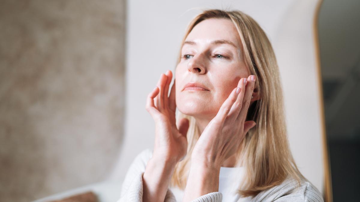 A focused adult woman practices facial self-massage in her well-lit bathroom. Using her fingertips, she gently smoothes her skin to improve circulation, reduce tension, and promote a healthy, natural glow. This authentic at-home wellness moment captures the popular beauty and self-care trend