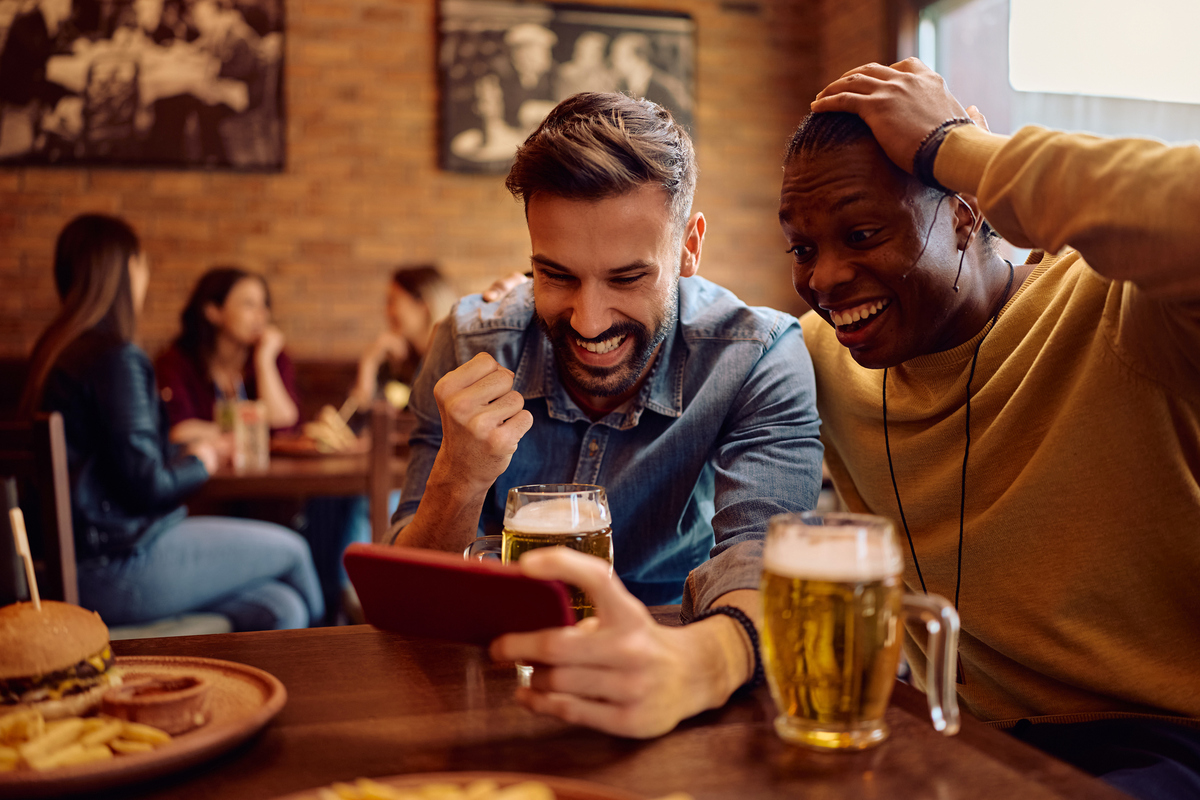  Happy male friends watching sports match on mobile phone while drinking beer in a pub. (Drazen Zigic/Getty) 